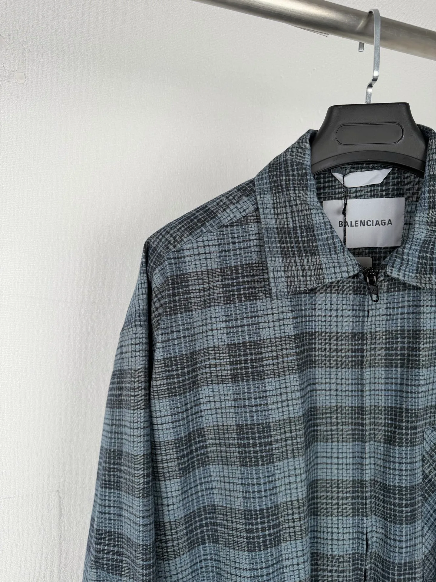 BALENCIAGA/balenciaga blue and gray plaid zipper pocket logo quilted jacket combines a three-layer structure of plaid woven fabric, filling cotton, and lining, and integrates plaid alignment technology and quilted garment production standards. There are multiple process difficulties in the whole process. The surface layer of the body is made of blue-gray Retro plaid fabric, and the cutting machine production strictly follows the global grid standards. The arrangement direction of the cutting pieces is fixed and cannot be changed at will. The horizontal and vertical lines of shoulder seams, side seams, and zipper placket joints must be consistent and coherent. The left chest patch pocket is cut independently, and the bag check pattern must be aligned with the front reference pattern. Slight deviations will destroy the visual integrity and directly increase fabric loss and cutting time. The most difficult part of the style is the three-layer composite sewing. The outer plaid fabric, filling cotton, and lining fabric have different physical properties, and the preshrinking processing parameters need to be adjusted separately. Pressure and temperature control are crucial during lamination operations. Once the parameters are out of balance, it can easily cause fabric stretch and distortion, plaid dislocation, and wrinkles and bulges in garments. At the same time, it is necessary to reasonably plan the quilting line to avoid the visual beauty of the grid pattern from being cut by the lines on the basis of firmly fixing the cotton layer and preventing cotton from running away. The placket adopts a centered zipper structure. It is difficult to sew the zipper tape and the quilted panels. It is required that the width of the left and right plackets be equal, the zipper routing is straight and smooth, and there is no lag in opening and closing. When the shirt lapel is filled with cotton, the thickness of the cotton needs to be controlled to prevent the collar from becoming bloated and unnaturally folded. The metal nameplate of the chest pocket is sewn at fixed points to avoid pulling the fabric during the operation and ensure that the plaid pattern is smooth and not deformed. The quilted material increases the overall thickness of the fabric. When sewing, you need to continuously adjust the presser foot pressure and stitch length to adapt to changes in sandwich thickness. The low-temperature and gentle ironing process is used for post-finishing to prevent the plaid from fading and the cotton layer from clumping due to high temperatures. From the precise alignment of cutting pieces, three-layer fabric pretreatment, composite quilting to garment finalization inspection, the processes are interlocking and the error tolerance rate is extremely low, testing the accuracy of plate making, equipment control and turning technology. Size:XS S M L